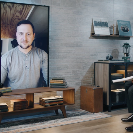 Man in grey shirt appears on television screen while being interviewed by a man in another room, wearing a suit.