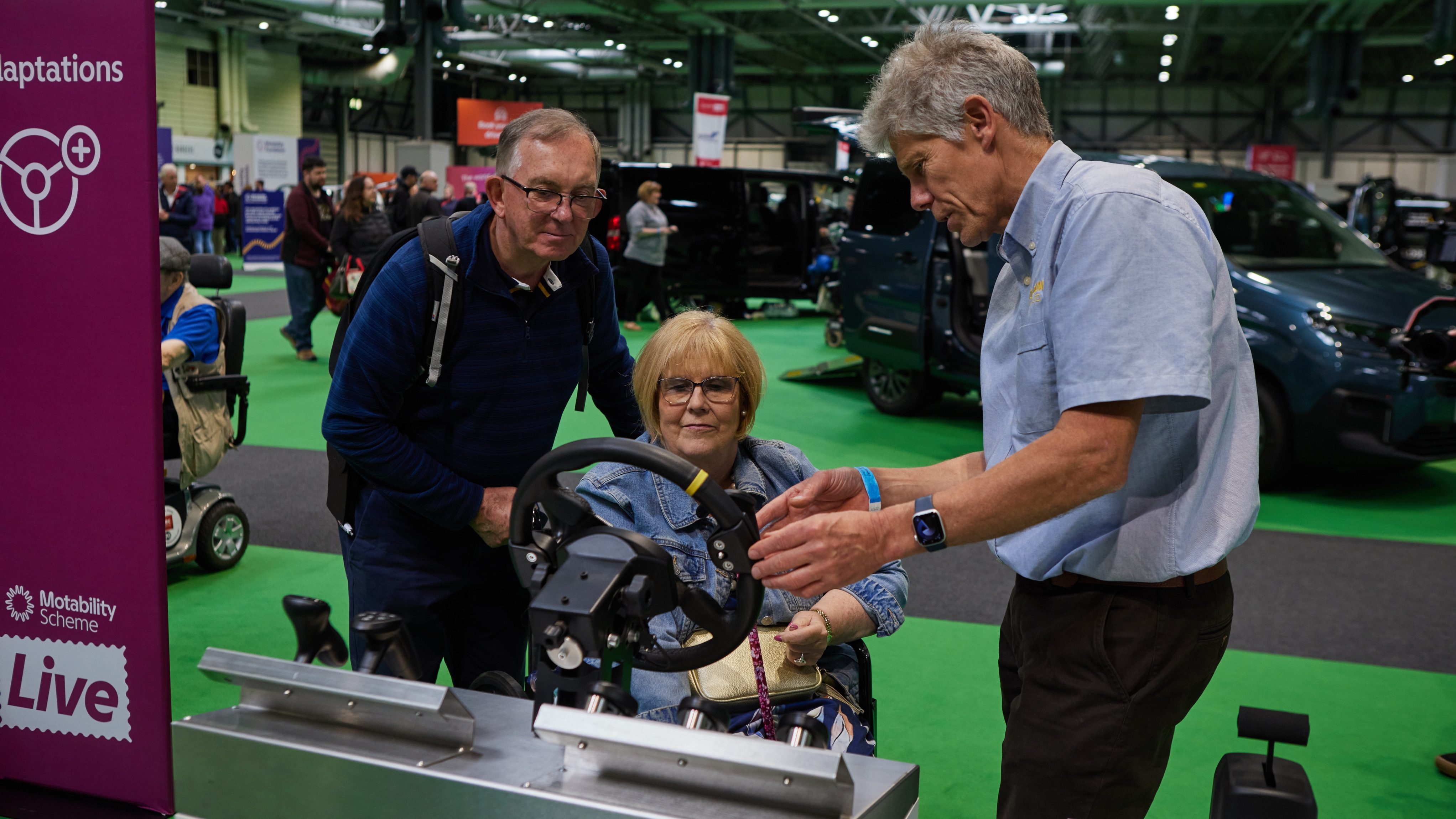 A woman uses a driving simulator while two men guide her at Motability Scheme Live in Birmingham. Accessible vehicles and displays are visible in the background.