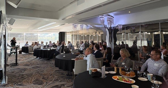 A large, bright event room with round tables where people are seated, listening to a speaker at a podium.