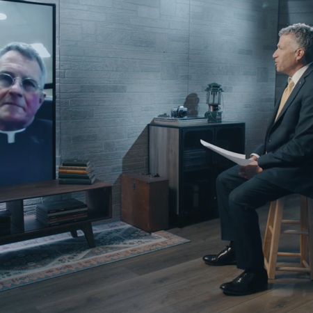 Catholic priest appears on a television while being interviewed by another man sitting on a stool.