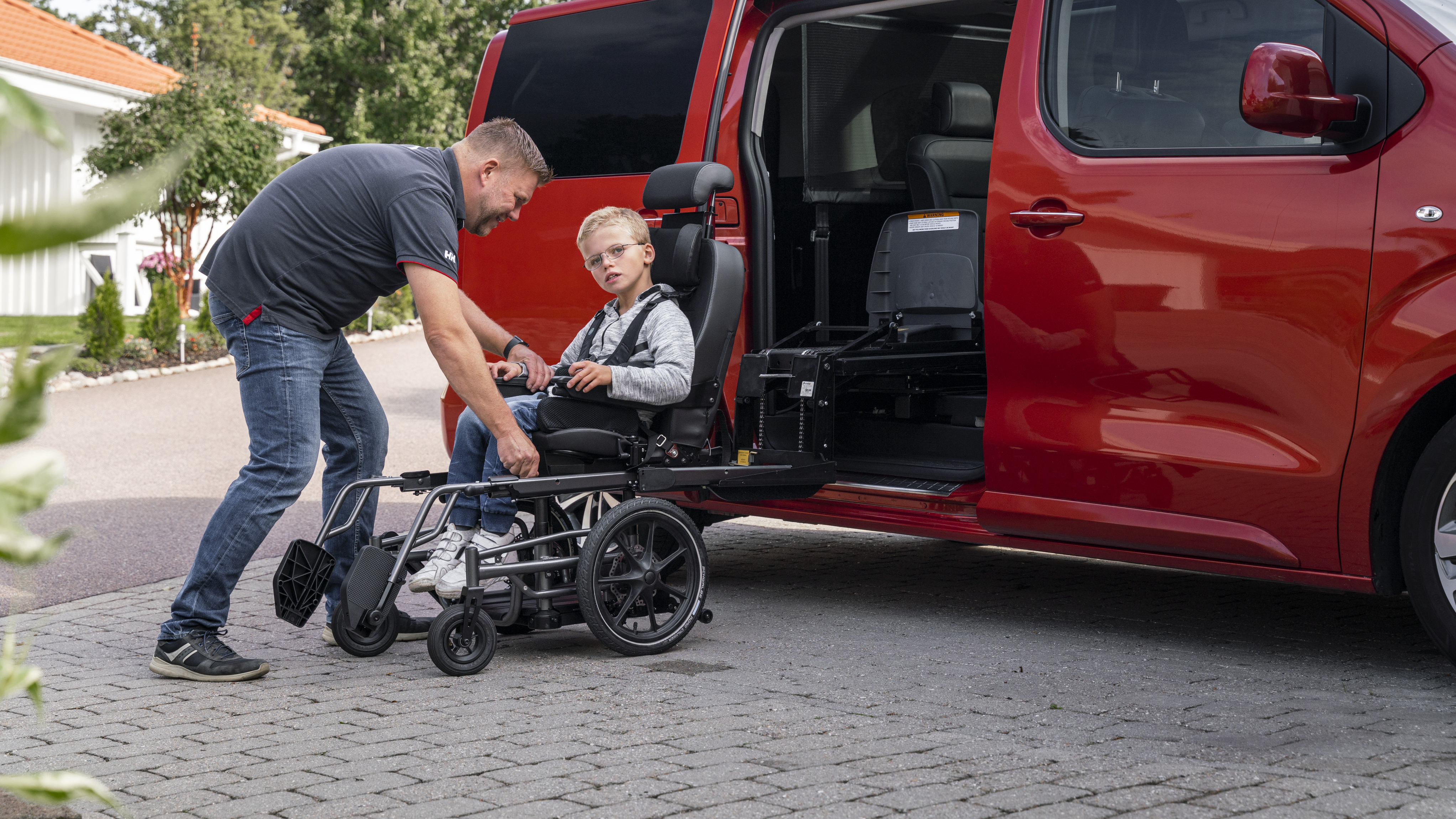 A man helps a young boy in a wheelchair transfer into a red accessible vehicle using a Carony system.