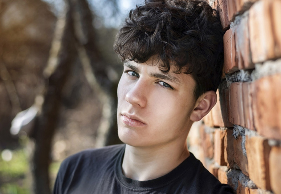 A young man with curly dark hair leans against a red brick wall outdoors, looking at the camera with a serious expression. He is wearing a black t-shirt and is surrounded by natural light.