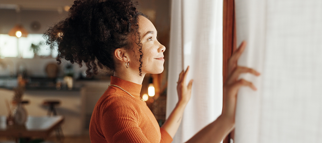 A Black woman stands in her home smiling out the window