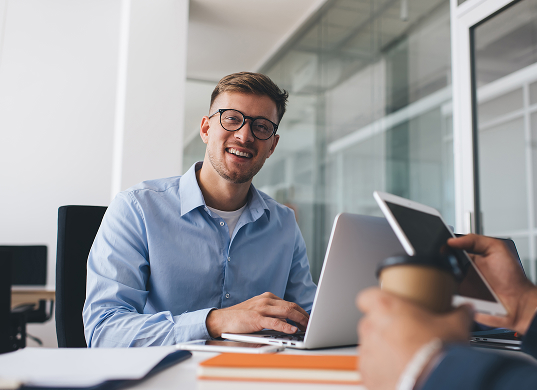 A young man with glasses and a light blue shirt smiles directly at the camera while sitting at a desk with a laptop open. Part of another person's hand holding a takeaway coffee cup is visible in the foreground.
