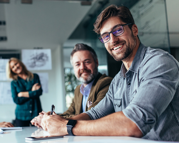 A man with dark-rimmed glasses and a grey shirt smiles directly at the camera while sitting at a conference table with a pen in his hand. Two other people, a man with a beard and a woman with blonde hair, are smiling in the blurred background.