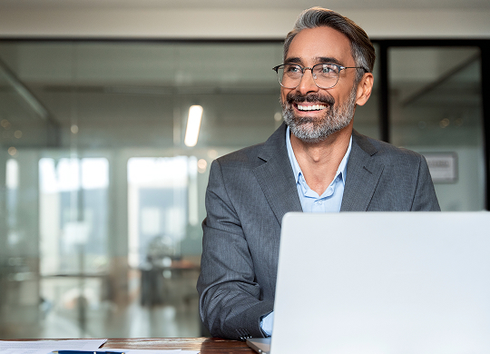 A smiling, middle-aged man with a gray beard and glasses, wearing a light blue shirt and a grey suit jacket, sits behind a silver laptop in an office. He is looking off to the side.