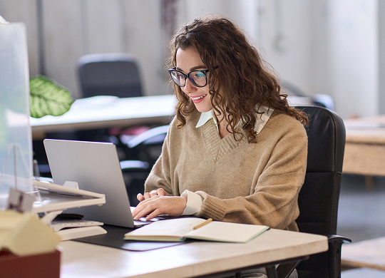 A smiling young woman with glasses and curly brown hair sits at a desk, typing on a silver laptop. She wears a beige V-neck sweater over a white collared shirt. A notebook and pen are on the desk in front of her.