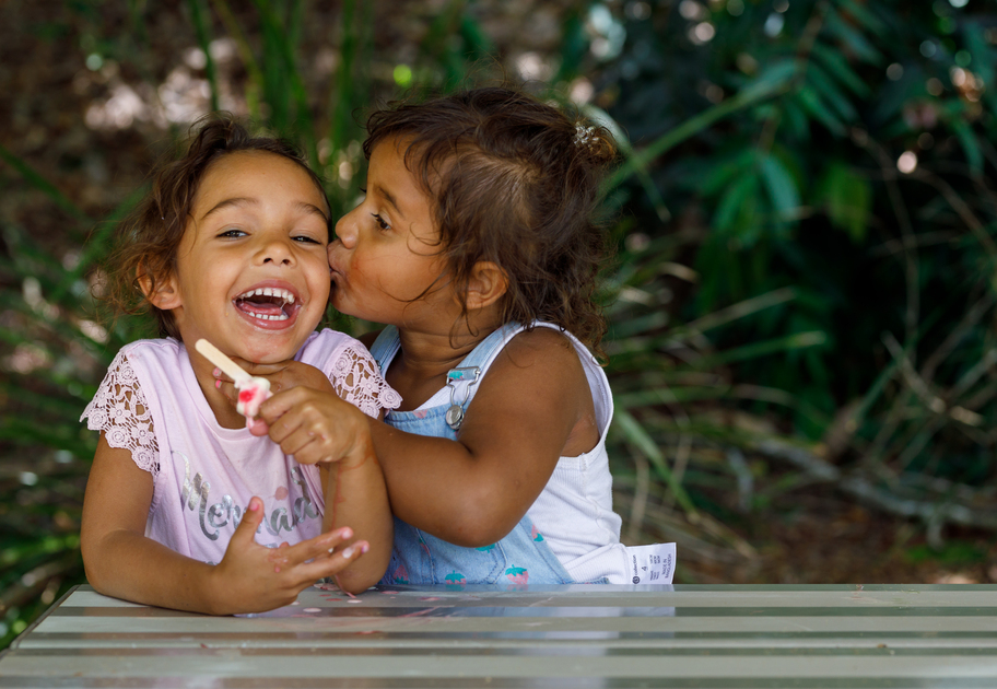 Two young girls sit outside on a bench. One is kissing the other on the cheek as the other playfully giggles.