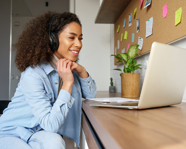 A young woman with curly hair, wearing a denim shirt and black over-ear headphones, smiles while looking at a laptop screen on a wooden desk. A corkboard and houseplant are visible behind her.