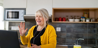 A smiling older woman in a yellow cardigan uses her laptop and earphones for an online language learning class in her kitchen.