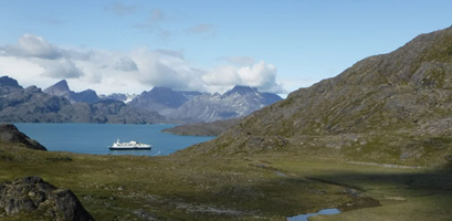 A small ship sails on the water through a valley of green, rolling hills, with a large, rocky mountain range in the background.