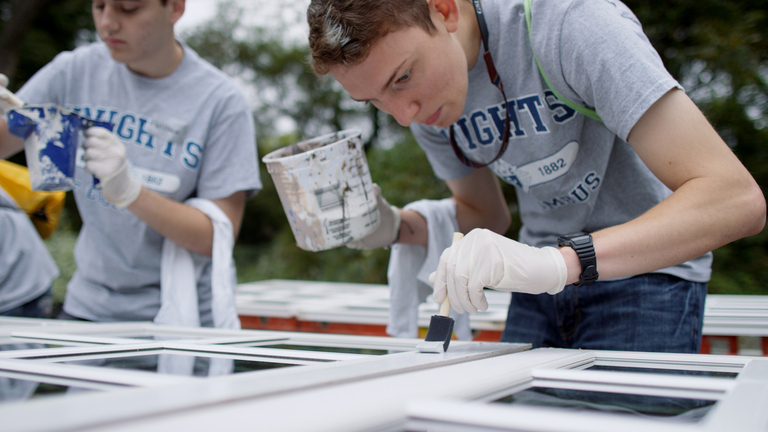 Photo of two college men in gray Knights of Columbus t-shirts painting windows with white paint.