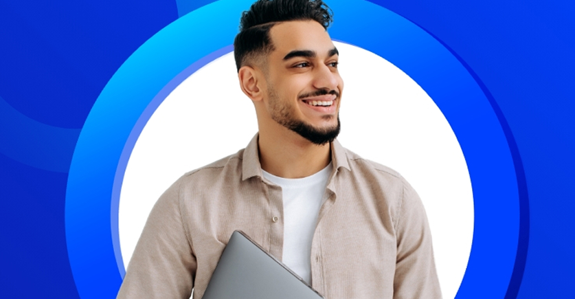 The image is a head-and-shoulders portrait of a smiling man. He has a beard and short, styled dark hair. He is wearing a light beige or tan collared shirt over a white t-shirt and is holding a closed silver laptop. He is looking off to the side with a happy and positive expression.