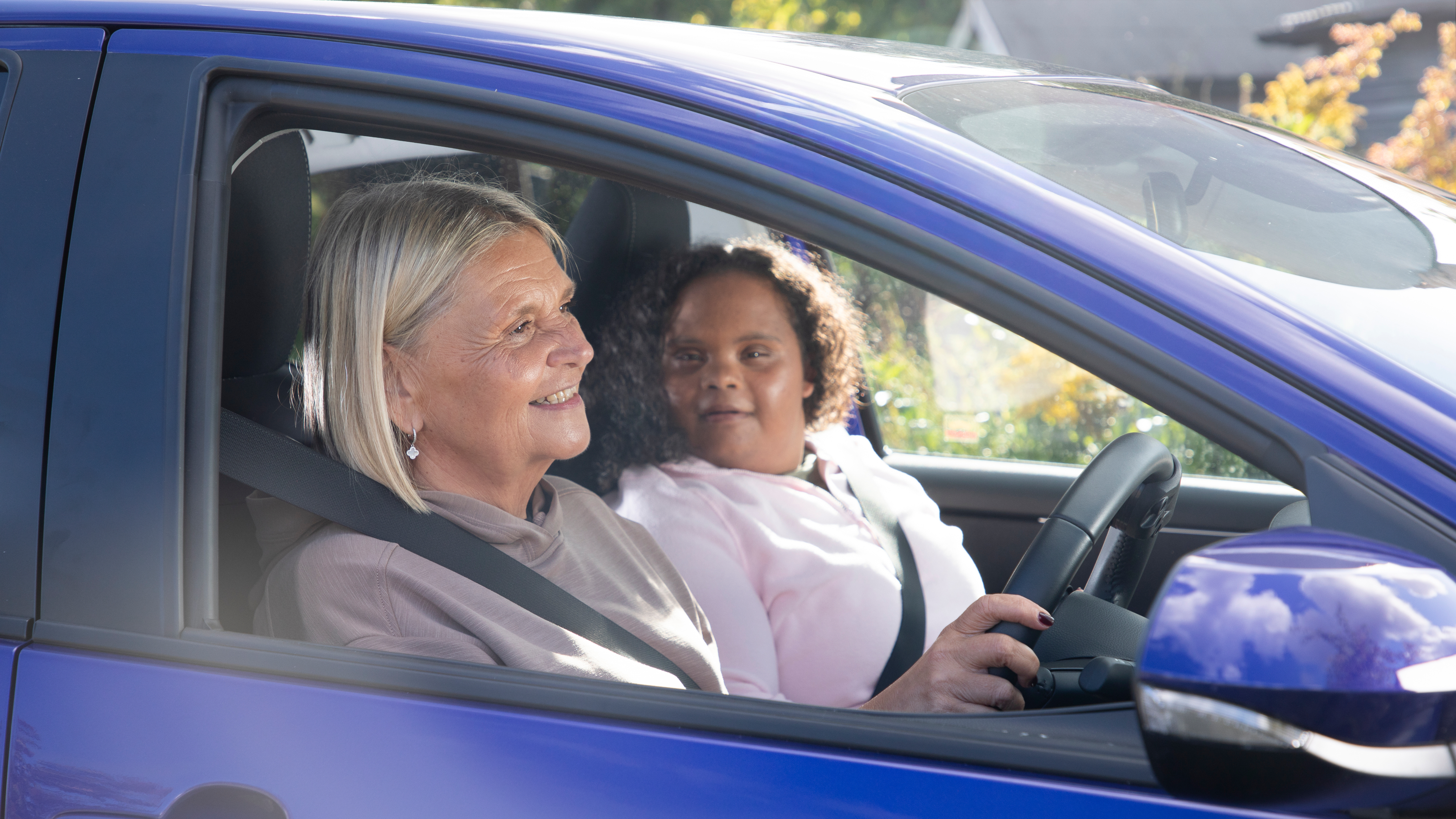 Two women sit in the front of a blue car, with the driver smiling while the passenger looks towards her, both wearing seatbelts in a bright, residential setting.