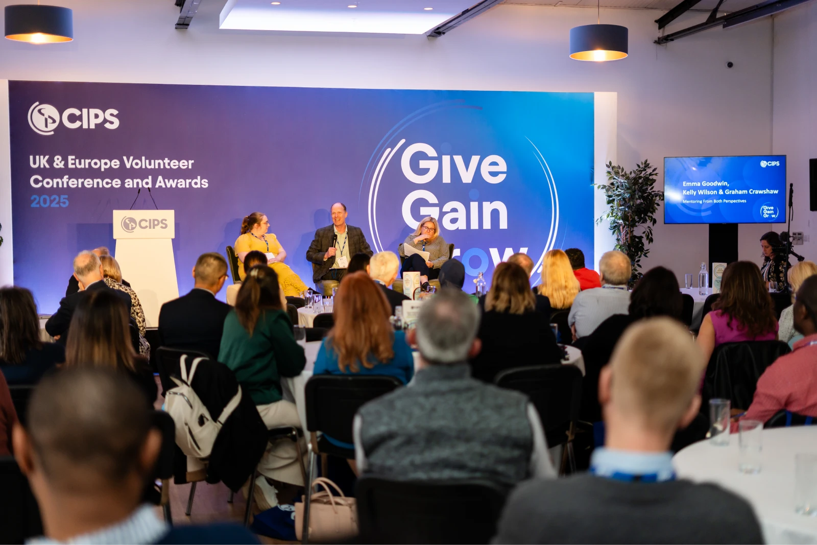 Panel discussion with three speakers seated on stage in front of a large CIPS UK & Europe Volunteer Conference and Awards 2025 banner that reads 'Give Gain Grow.' An audience is seated at tables watching the stage.