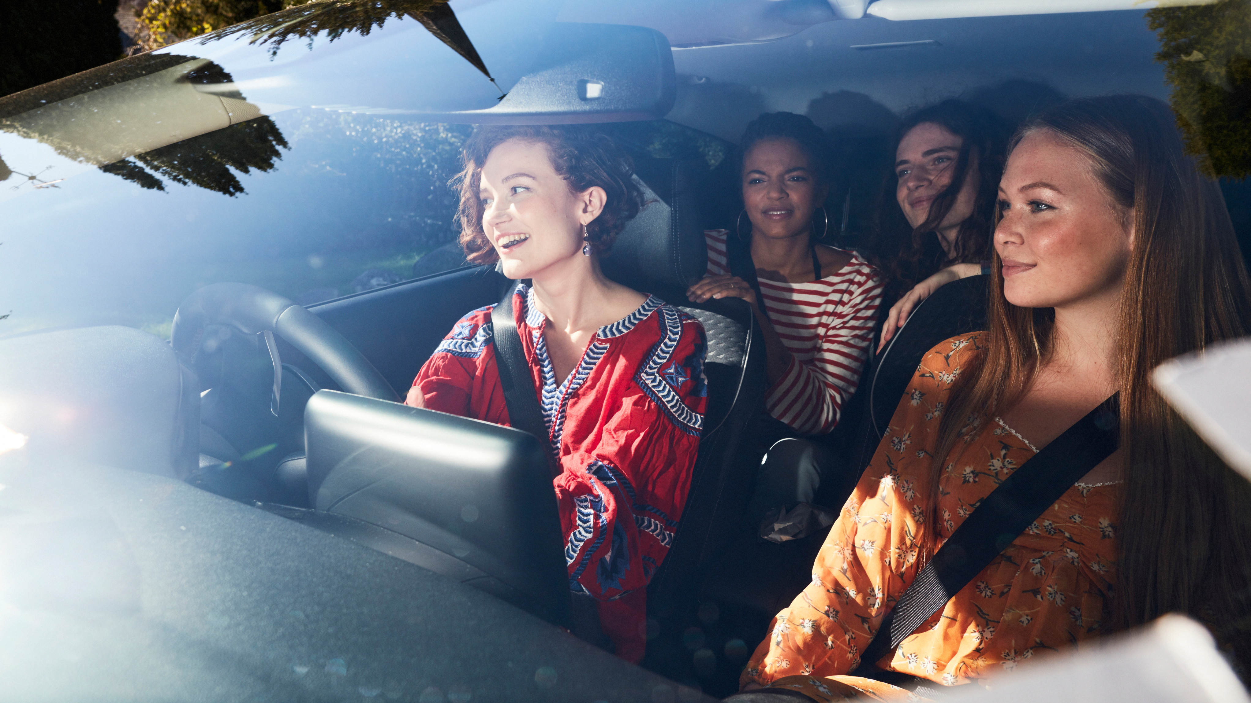 Four women sit inside a car, with one driving and three passengers smiling and chatting, sunlight streaming through the windscreen during a daytime journey.