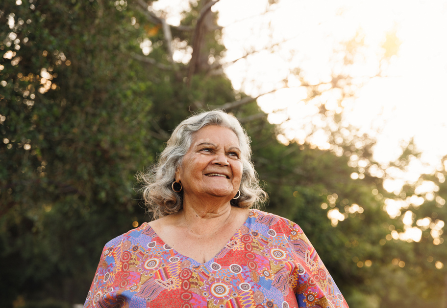 Older woman with gray hair and hoop earrings smiling outdoors, wearing a colorful patterned top. She stands in front of green trees with sunlight filtering through the branches in the background.