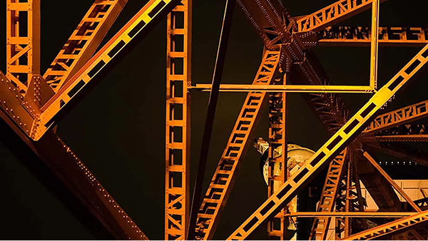 A close-up view of the intricate, bright orange, crisscrossed steel framework of a large structure against a dark night sky.