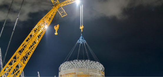 A large yellow construction crane, brightly illuminated, lifting a circular structural piece at a construction site against a dark night sky.