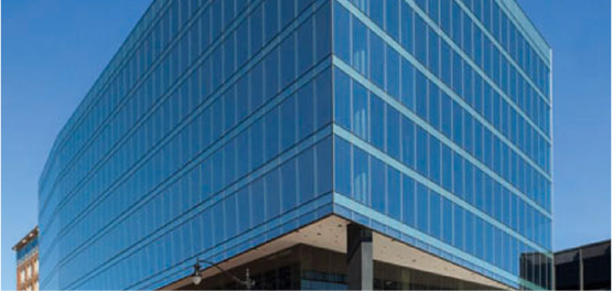 A modern, multi-story office building with a glass curtain wall reflecting the blue sky, seen from a street-level perspective.