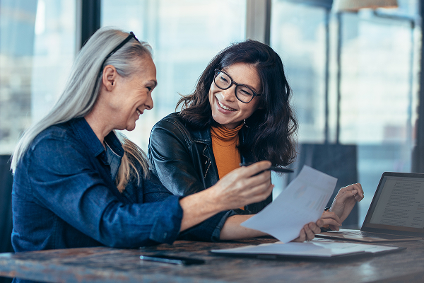 Two smiling women are seated together indoors, one with grey hair and a denim shirt, the other with dark hair, a leather jacket, and an orange top. They are looking down at a document, with one woman pointing to it, suggesting collaboration or review.