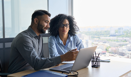 A man and a woman seated side-by-side at a desk in a modern high-rise office with large windows overlooking a city skyline. The man is pointing at a laptop screen while the woman looks on, suggesting a discussion or instruction.