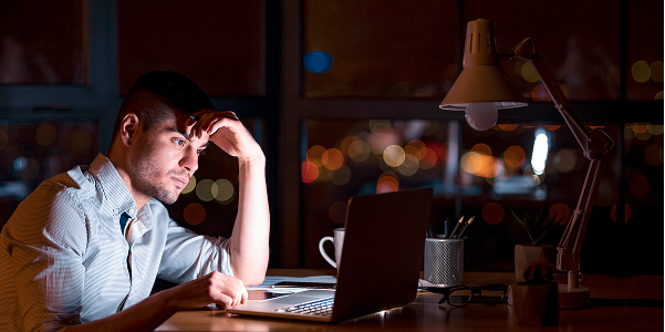 A focused man sits at a desk late at night, working on a laptop with his hand resting on his forehead, illuminated by a desk lamp against a blurred background of city lights.