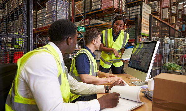 Three logistics or warehouse workers in high-visibility vests look at a computer screen and paperwork at a desk in a busy warehouse.