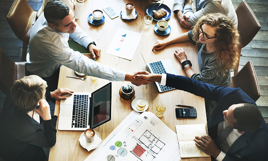 An overhead view of a diverse group of six professionals seated around a conference table. Two of the individuals are shaking hands over the table, with laptops, coffee cups, pastries, and business documents scattered around.