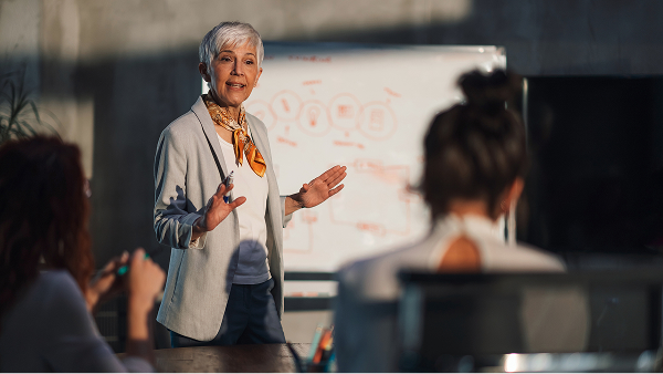 A mature woman with short white hair, wearing a blazer and a patterned scarf, is giving a presentation and gesturing at a whiteboard to an audience in a dim room.