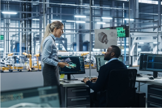 A woman holds a tablet while talking to a man seated at a desk with two computer screens displaying engineering designs, inside a modern manufacturing facility.