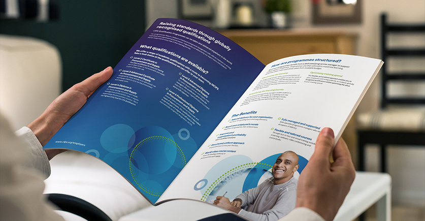 A person holds an open brochure or booklet titled "Raising standards through globally recognised qualifications," showing a blue and white design with text and a small photo of a smiling man.