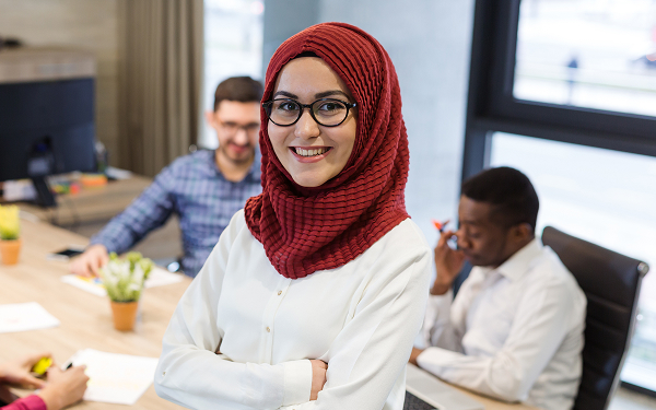 A young woman wearing glasses and a red hijab stands in a modern office, smiling at the camera with her arms crossed. Two coworkers are seated at a conference table behind her.