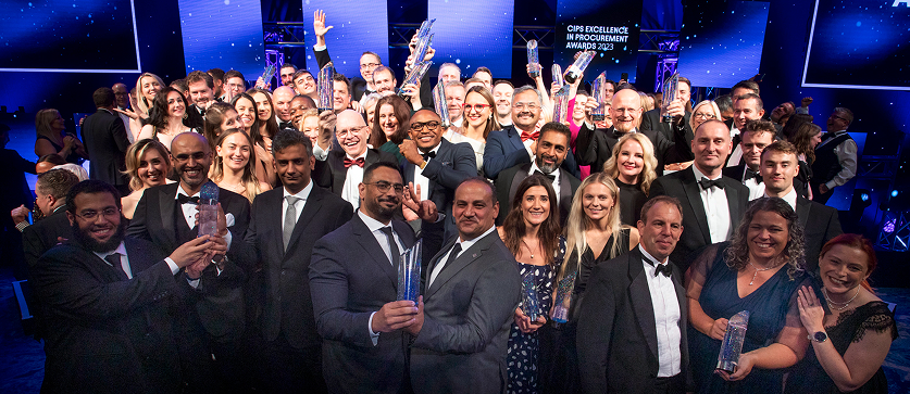 A large group of people in formal attire (suits, tuxedos, and gowns) are posing together on a stage, cheering and holding various trophies or awards. A screen in the background reads "CIPS EXCELLENCE IN PROCUREMENT AWARDS 2023".