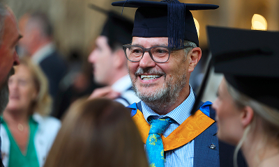 A smiling graduate in a navy suit, blue and yellow academic hood, and black mortarboard cap is talking to other attendees at a graduation ceremony. He is wearing glasses and a bright blue and yellow tie.