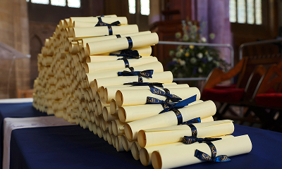 A large stack of rolled-up diplomas or certificates, tied with dark blue ribbons, is placed on a table covered with a dark blue cloth inside a graduation hall or church.