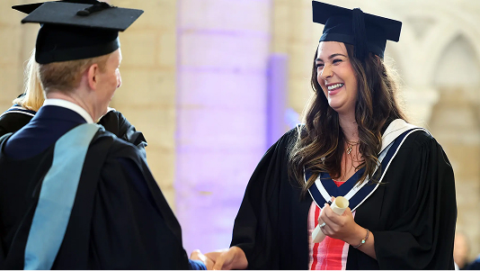A young woman in a graduation cap and gown, holding her rolled-up certificate, is smiling and shaking hands with another graduate in a black and blue gown during a ceremony in a large hall.