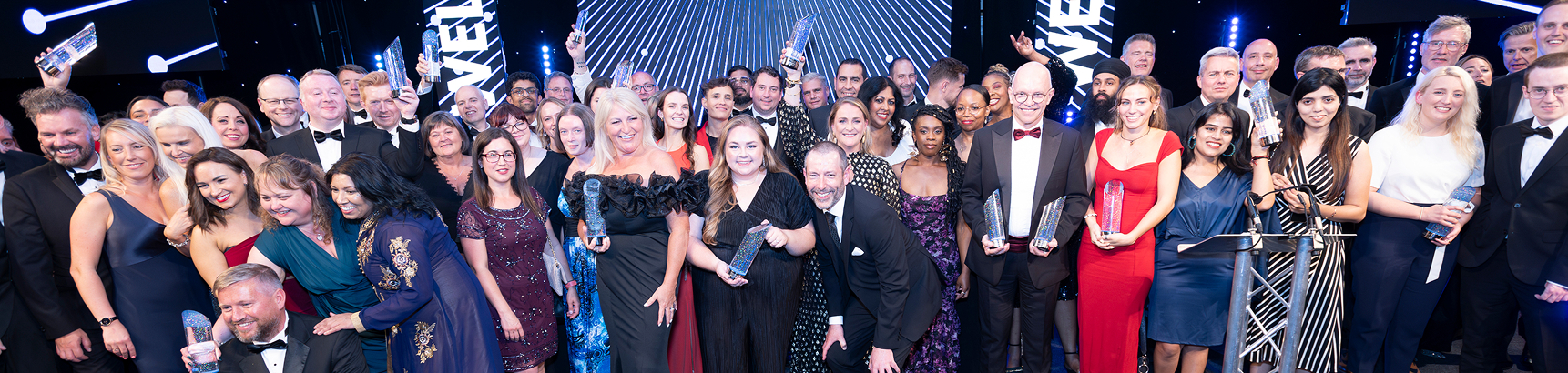 A large group of professionally dressed people, many holding awards or trophies, pose for a panoramic photo on a brightly lit stage at a formal awards event. They are smiling and cheering.