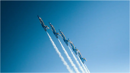 A line of four military-style jet aircraft flying in tight formation, climbing against a clear blue sky, leaving white contrails behind them.