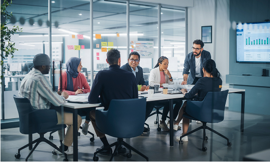 A diverse group of seven professionals sitting around a conference table in a modern glass-walled office, engaged in a discussion. One man is standing and presenting or directing the conversation.