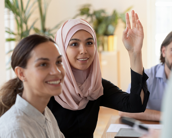 A smiling young woman wearing a pink hijab and a black top raises her hand to ask a question or answer one during a meeting or class. A second woman, out of focus, smiles in the foreground. A man's face is partially visible on the right.