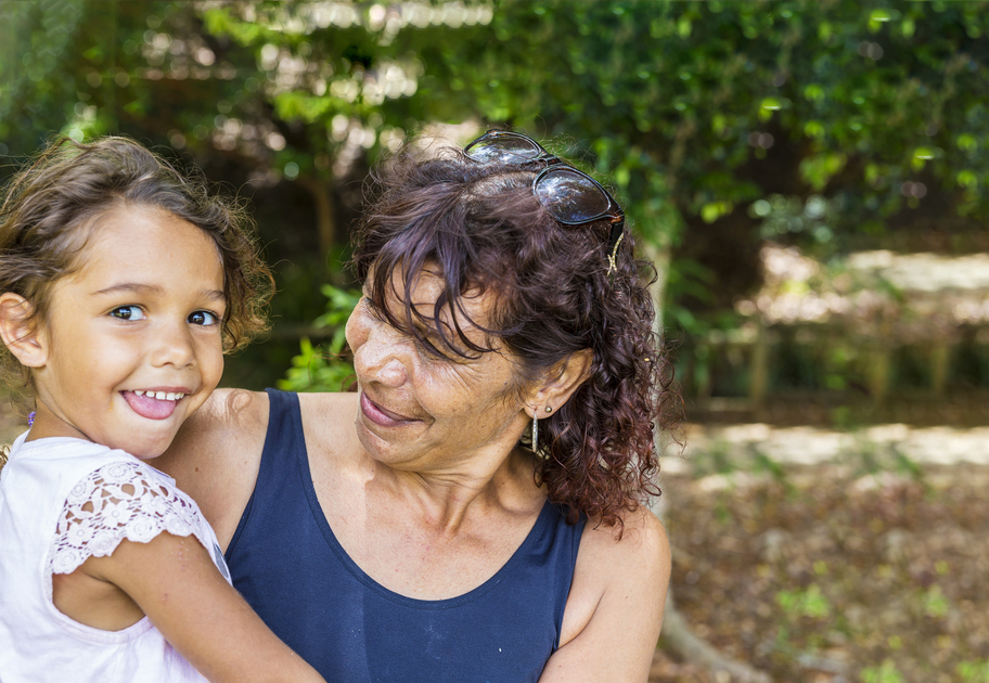 A lady with curly brown hair holds a young girl with a cheeky grin. Both are happy and are outside.