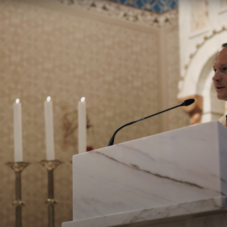 Catholic Priest stands at podium with microphone surrounded by candles