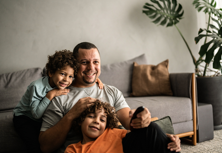 A man sits on a sofa smiling, holding a TV remote, with two young children hugging him happily. There are pillows and a plant in the background, creating a cozy living room scene.