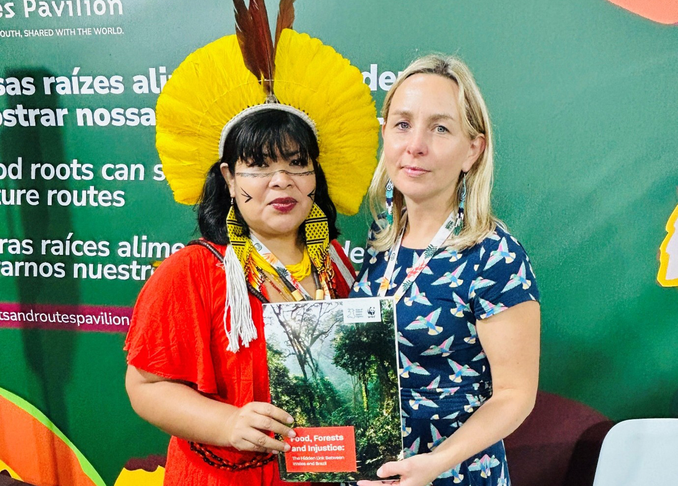 Two women stand together holding a book titled 'Food, Forests and Injustice' in front of a green display with multilingual text.