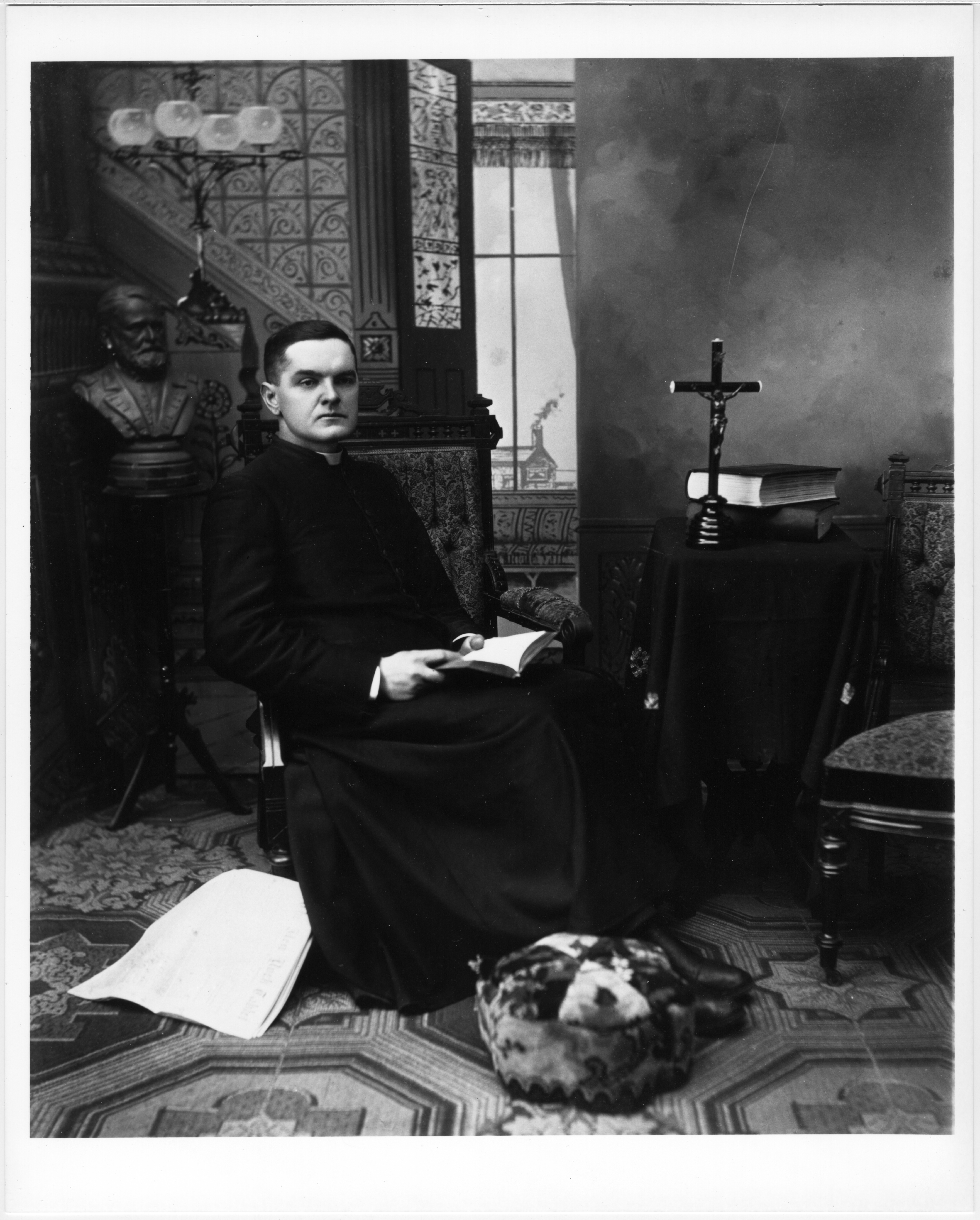 Father Michael J. McGivney seated in studio rectory setting, dressed in a long cassock, seated in high-backed upholstered chair, opened book in lap.