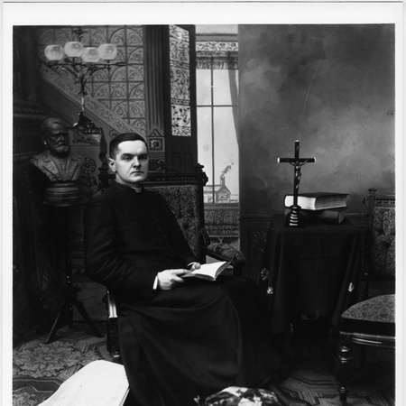 Father Michael J. McGivney seated in studio rectory setting, dressed in a long cassock, seated in high-backed upholstered chair, opened book in lap.