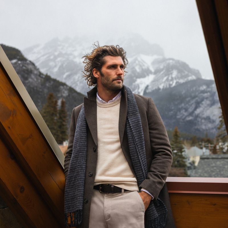 Person wearing a brown overcoat, beige knit jumper, trousers and a dark scarf, standing on a timber balcony with a mountainous, snow-covered landscape in the background.