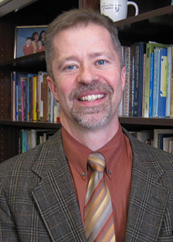 Headshot of Moral Theologian Dr. John Grabowski, who smiles while wearing a tan plaid suit, and a striped tie