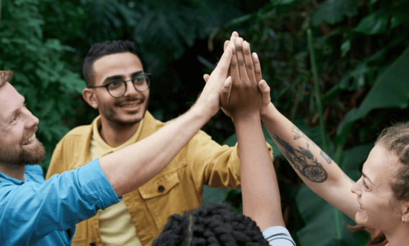 A group of four people standing outdoors in a lush green environment, giving each other a high-five. One person is wearing glasses and a yellow jacket, another has tattoos on the arm, and the others are partially visible.